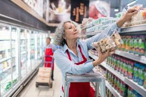 Older woman store worker restocking items on a shelf while male store worker pushes dolly cart with boxes next to freezer section